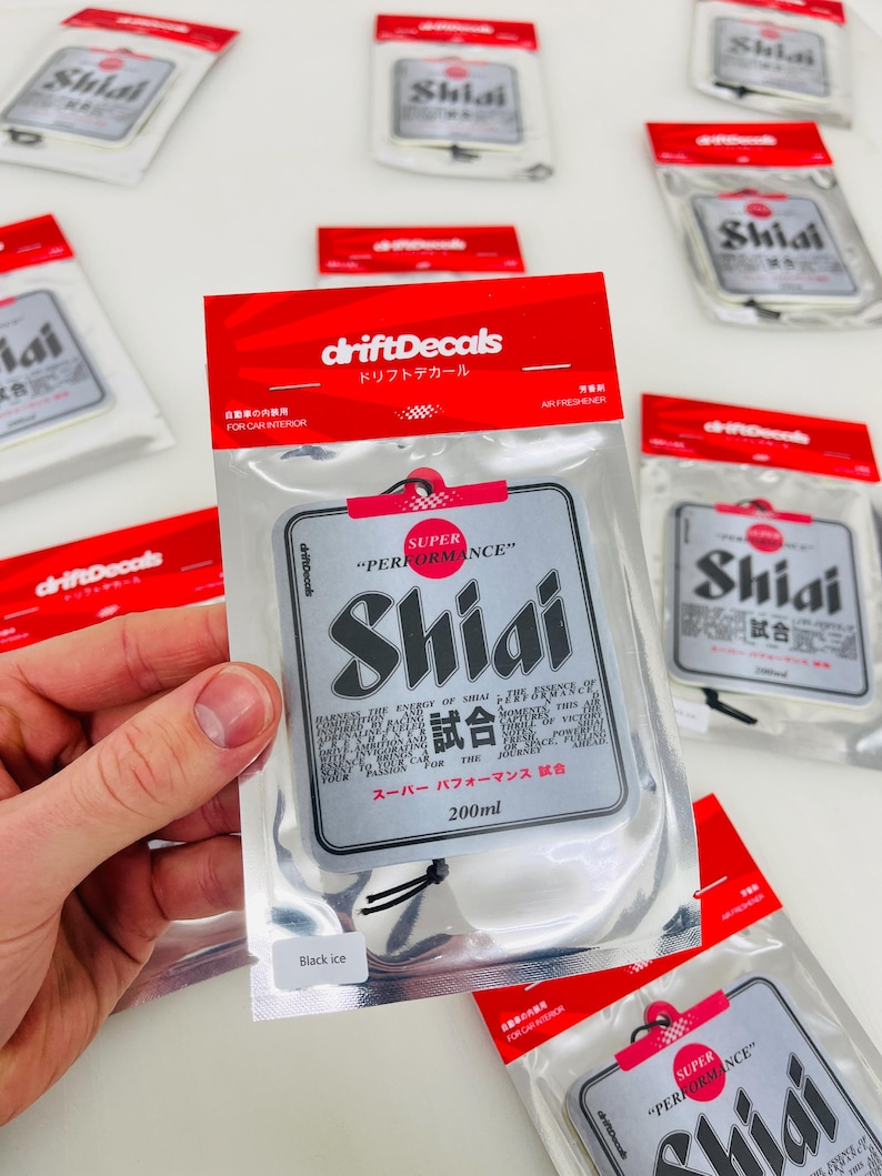 May include: A silver car air freshener with the text "Shiai" and "Super Performance" in black and red. The air freshener is packaged in a clear plastic bag with a black string.