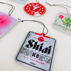 May include: A silver car air freshener with the text "SUPER PERFORMANCE" and "Shiai 試合" in black and red. The text "200ml" is printed in black at the bottom of the air freshener.