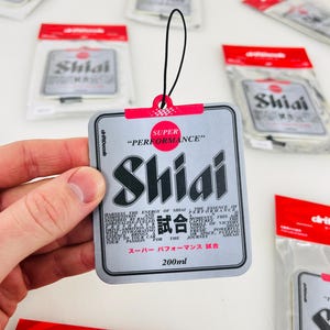 May include: A silver car air freshener with the text "SUPER PERFORMANCE" and "Shiai" in large black letters. The text "試合" is in smaller black letters. The air freshener is 200 ml and has a black string for hanging.