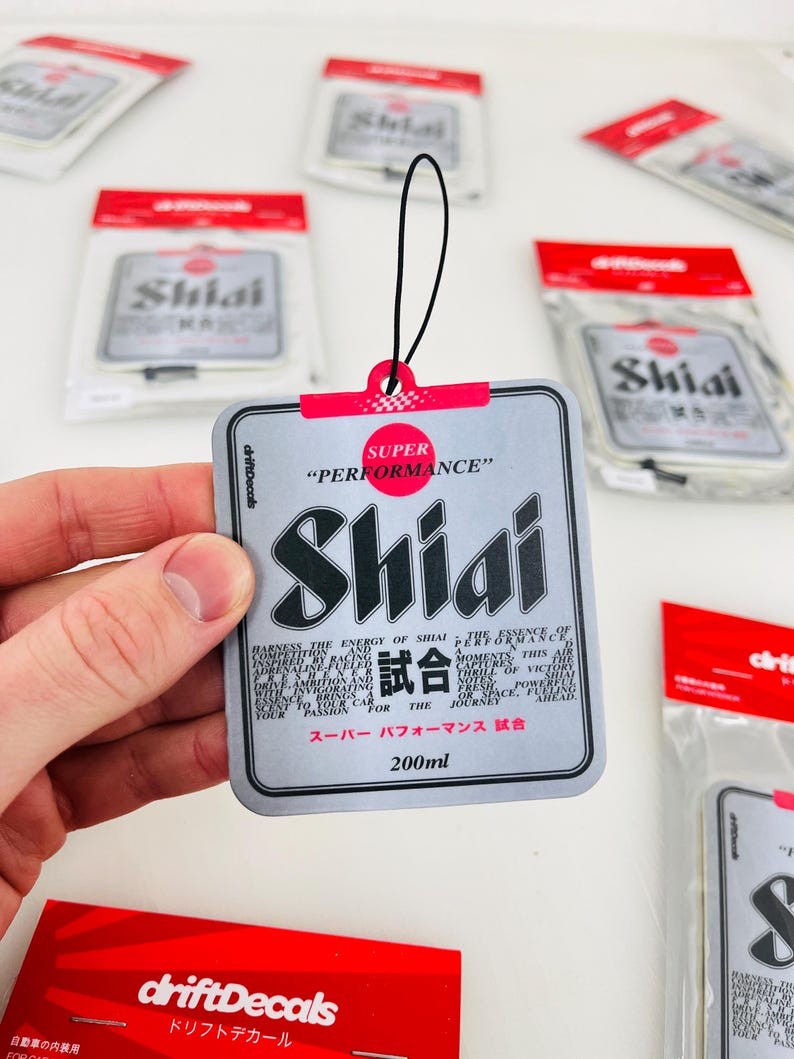 May include: A silver car air freshener with the text "SUPER PERFORMANCE" and "Shiai" in large black letters. The text "試合" is in smaller black letters. The air freshener is 200 ml and has a black string for hanging.