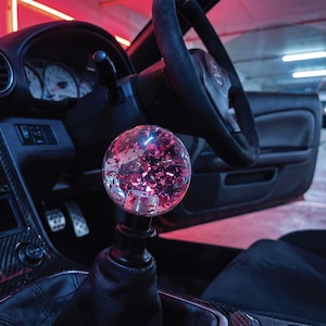 May include: A spherical, clear gear shift knob featuring embedded pink and red metallic flakes, mounted on a black base within a vehicle interior. The knob is positioned in the centre console, surrounded by a black dashboard, steering wheel, and gear boot.