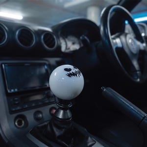 May include: A spherical white gear shift knob featuring a black graphic of a grinning face with stitched eyes and mouth. The knob is mounted on a black metal base with a black leather boot, positioned in the centre console of a vehicle interior.
