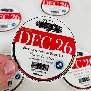 Può includere: Adesivo circolare con grafica di auto nera e la scritta "DEC 26" in rosso. L'adesivo include anche il testo "Bayerische Motoren Werke A.G" e un codice a barre. Il logo BMW è visibile.