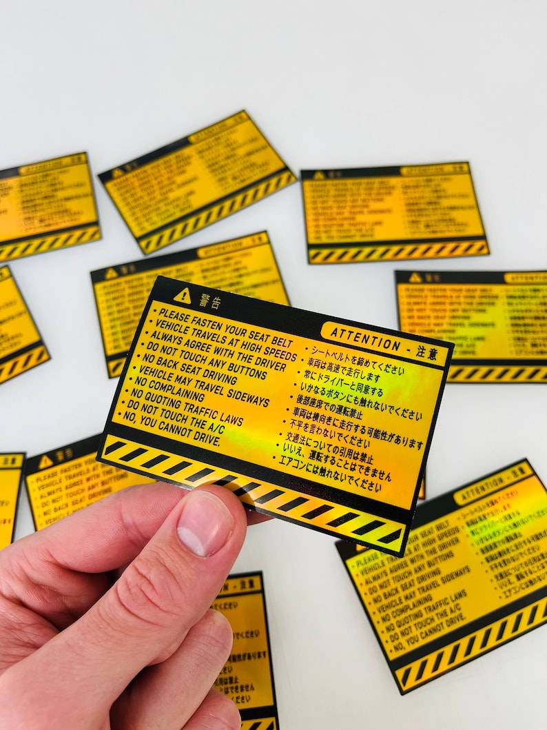 May include: A yellow and black warning sticker with a black and white graphic of a warning sign. The sticker has text in both English and Japanese. The text reads: "Please fasten your seat belt. Vehicle travels at high speeds. Always agree with the driver. Do not touch any buttons. No back seat driving. Vehicle may travel sideways. No complaining. No quoting traffic laws. Do not touch the A/C. No, you cannot drive."