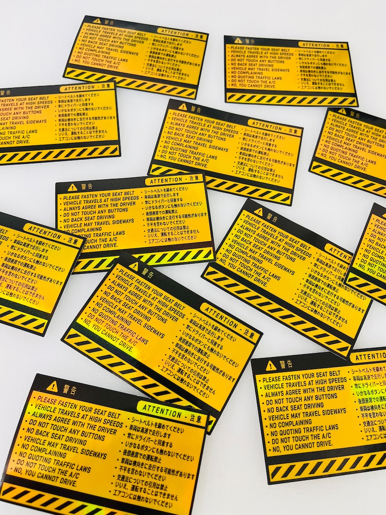 May include: A set of ten yellow and black warning stickers with the text "ATTENTION - 注意" and a list of safety instructions in Japanese and English. The instructions include "Please fasten your seat belt", "Vehicle travels at high speeds", "Always agree with the driver", "Do not touch any buttons", "No back seat driving", "Vehicle may travel sideways", "No complaining", "No quoting traffic laws", "Do not touch the A/C", and "No, you cannot drive."