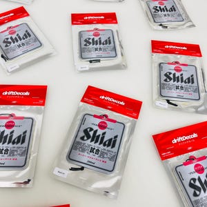 May include: A set of 10 silver car air fresheners with a black and white design. The air fresheners feature the text "Shiai" and "試合" in Japanese. The air fresheners are packaged in clear plastic bags with red and white labels that say "driftDecals" and "ドリフトデカール".