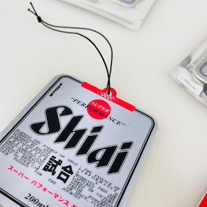 May include: A silver car air freshener with a black and white logo that says "Shiai" and the text "Super Performance" in red. The air freshener is shaped like a rectangle and has a black string for hanging. The text "試合" is also on the air freshener.