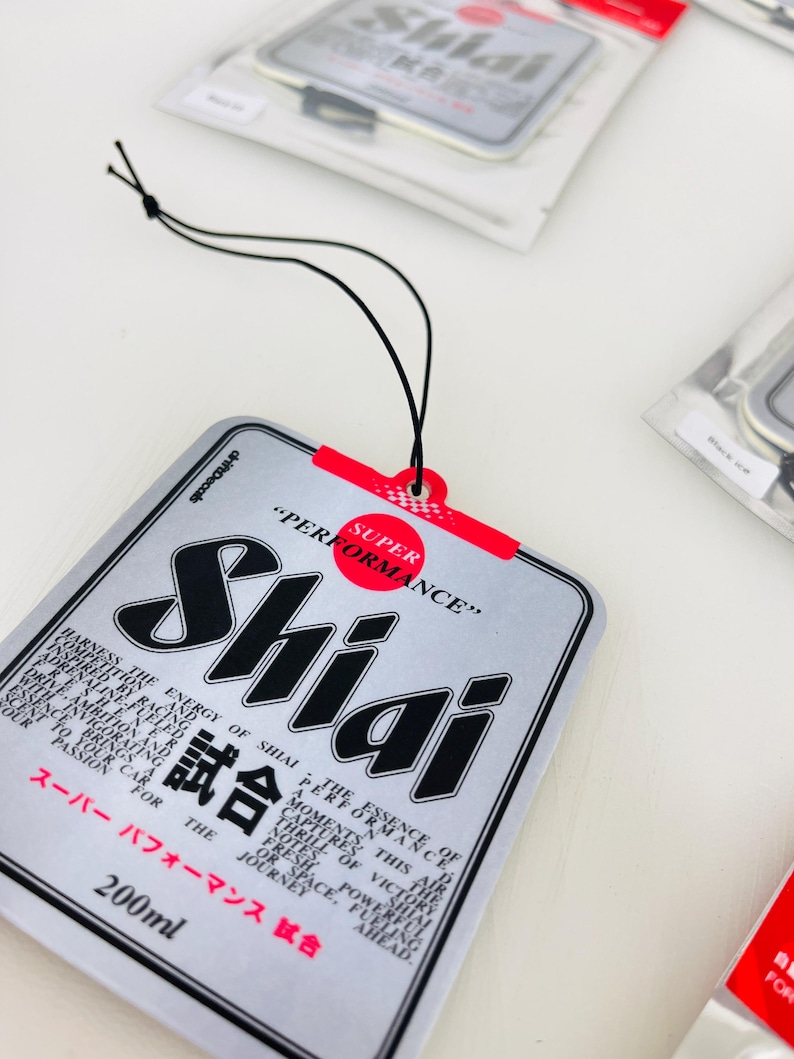 May include: A silver car air freshener with a black and white logo that says "Shiai" and the text "Super Performance" in red. The air freshener is shaped like a rectangle and has a black string for hanging. The text "試合" is also on the air freshener.