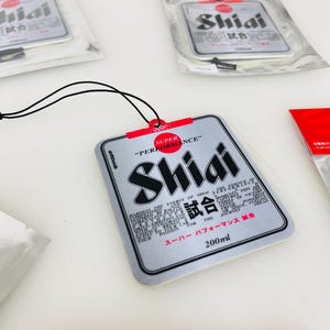 May include: A silver car air freshener with a black string for hanging. The air freshener is shaped like a can and has the text "Shiai 試合" and "Super Performance" printed on it. The air freshener is 200 ml.