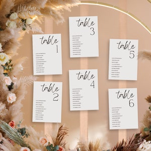 Könnte beinhalten: Sechs weiße Tischkarten mit schwarzem Text, jede mit der Aufschrift "Tisch" gefolgt von einer Zahl von eins bis sechs. Die Karten sind vor einem beigen Hintergrund angeordnet, umgeben von Blumenarrangements und Pampasgras.