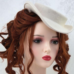 May include: A white felt hat with a wide brim and a slightly curved crown. The hat is worn on a mannequin with long, curly red hair.