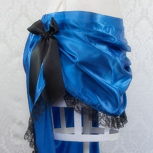 Op de afbeelding: Een helderblauwe rok met een zwarte strik en kanten rand. De rok is gedrapeerd en gerimpeld, met een high-low zoomlijn. De zwarte kanten rand voegt een delicaat contrast toe aan de blauwe stof. De rok wordt getoond op een witte paspop.
