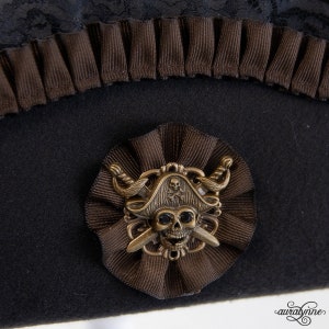 May include: Black pirate hat with brown accents. The hat features a decorative brown ribbon and a bronze-colored skull and crossbones emblem. A black lace trim is visible at the top of the hat.