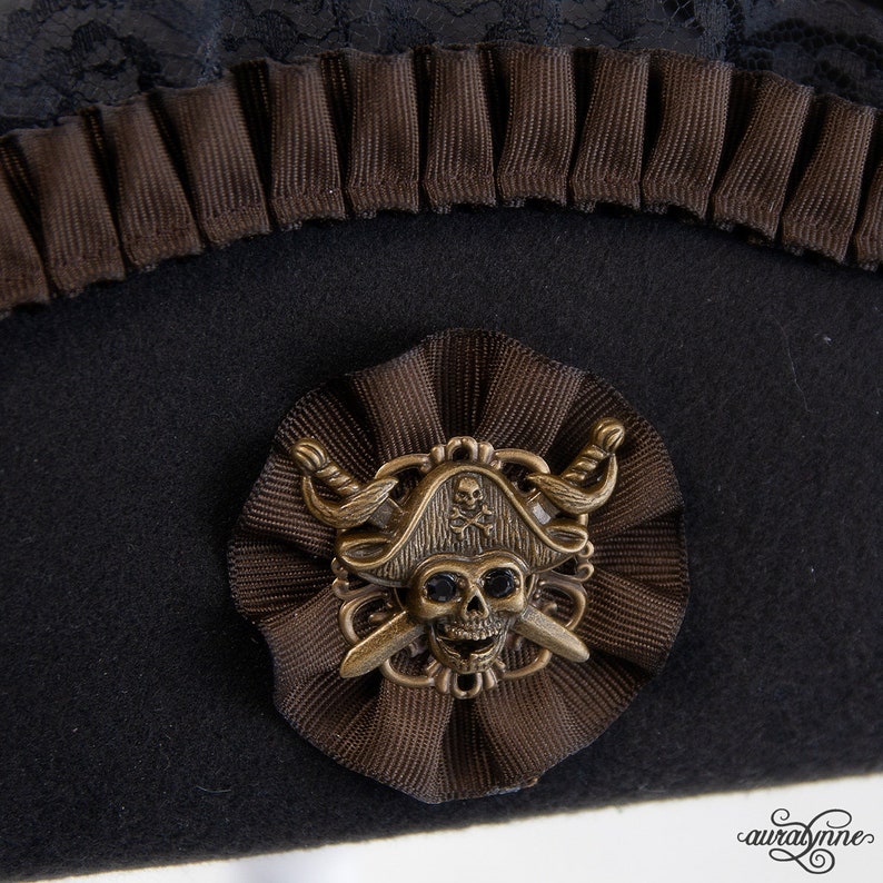 May include: Black pirate hat with brown accents. The hat features a decorative brown ribbon and a bronze-colored skull and crossbones emblem. A black lace trim is visible at the top of the hat.