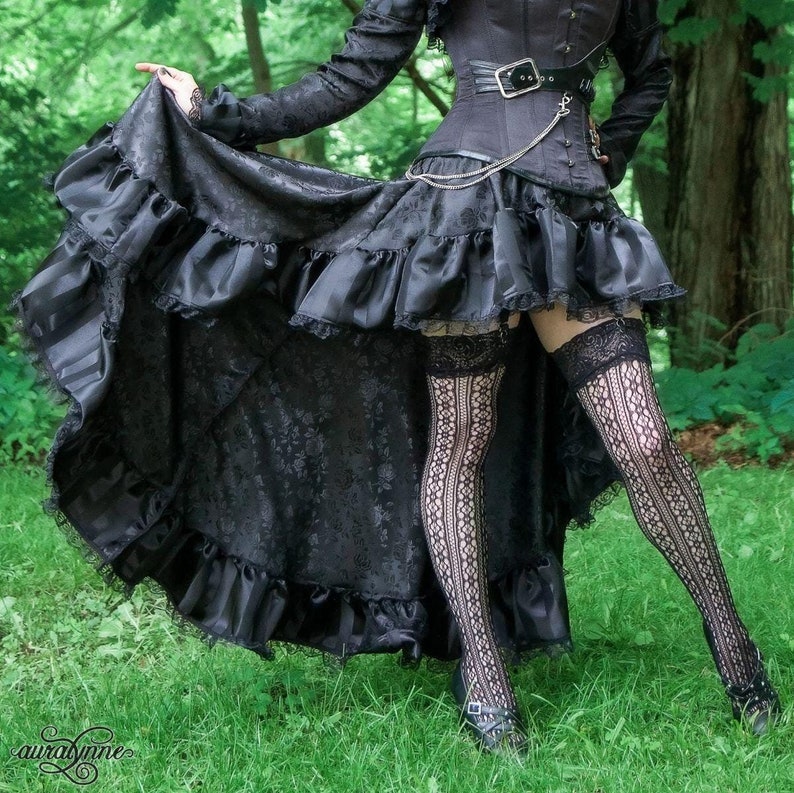 May include: A black, high-low skirt with a ruffled hem and lace trim. The skirt is made of a brocade fabric with a floral pattern. The skirt is paired with black stockings and black shoes.