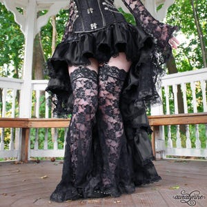 May include: A black corset top with silver chains and a high-low skirt featuring multiple ruffled layers. The outfit includes black lace thigh-high stockings with a floral pattern, worn with a long-sleeved black lace top with flared cuffs.
