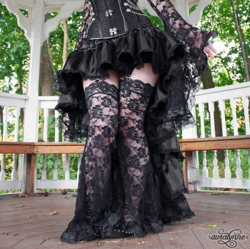 May include: A black corset top with silver chains and a high-low skirt featuring multiple ruffled layers. The outfit includes black lace thigh-high stockings with a floral pattern, worn with a long-sleeved black lace top with flared cuffs.