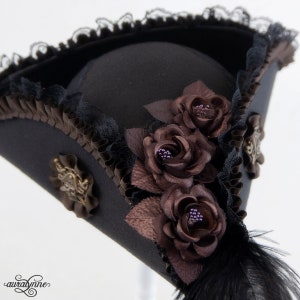 May include: Black tricorn hat with brown fabric roses and decorative accents. The hat features black lace trim, a black feather, and ornate bronze-colored embellishments. A stylish accessory for cosplay or themed events.