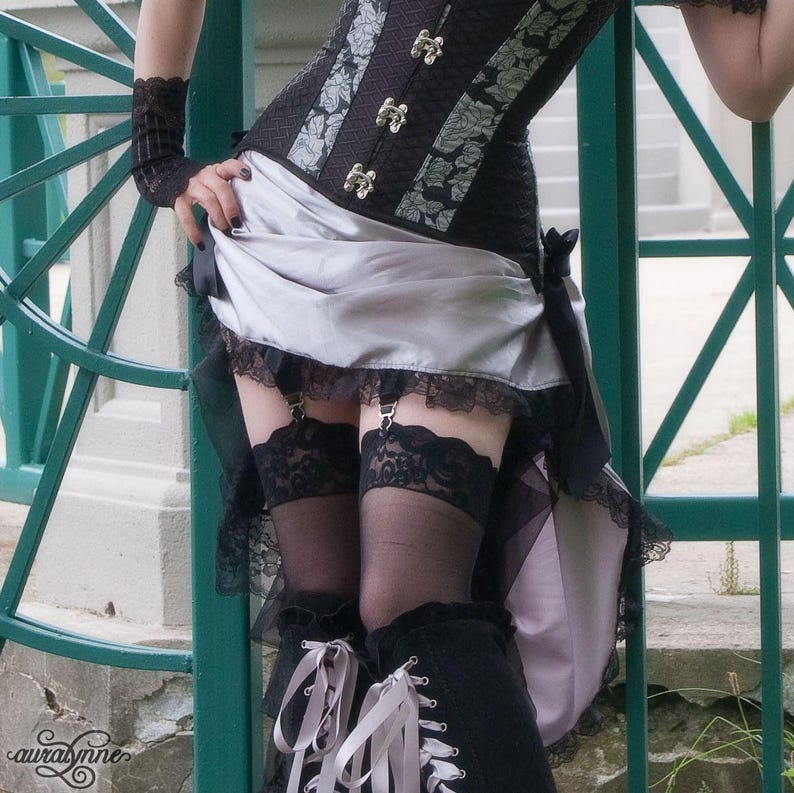 May include: A black corset with a silver floral pattern and silver metal clasps, paired with a silver skirt featuring black lace trim and black ribbon ties. The outfit includes black lace-topped stockings with garter clips and black lace wrist cuffs.