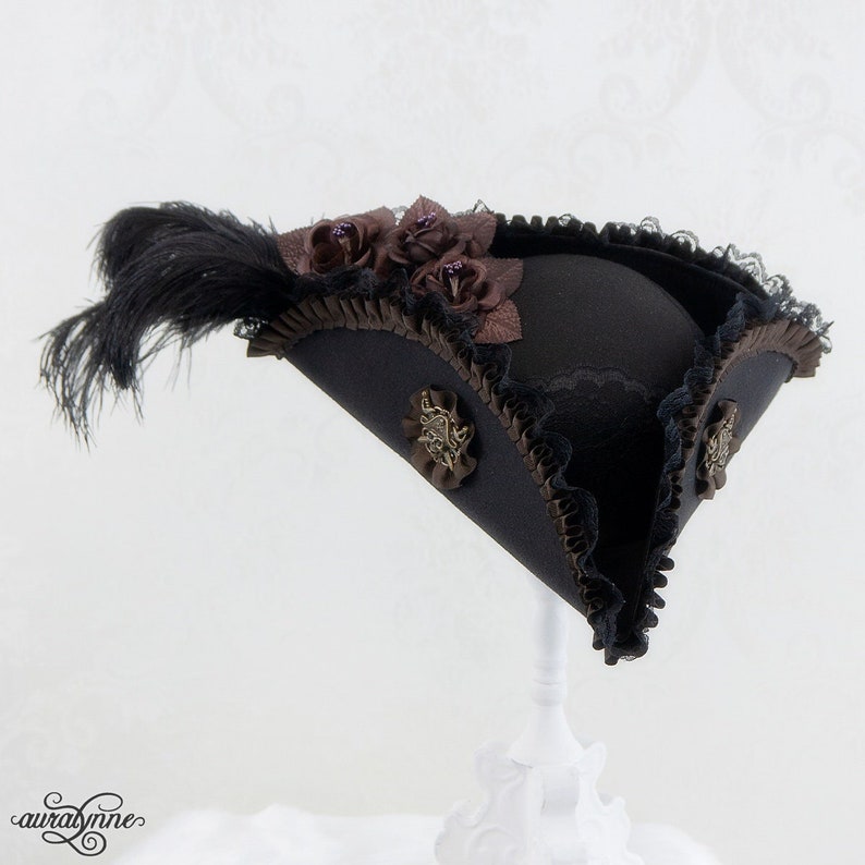 May include: A black tricorn hat with brown trim, lace, and decorative embellishments. The hat features a large black feather and brown fabric roses. The hat is displayed on a white stand.