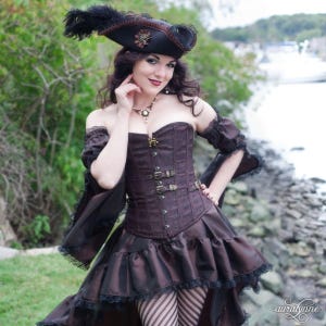 May include: A brown steampunk-style outfit featuring a corset with buckles, a ruffled skirt, and off-the-shoulder sleeves. The ensemble includes a black pirate hat with feathers and lace trim. A necklace and striped stockings complete the look.