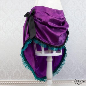 Pu&ograve; includere: Gonna in raso viola con design asimmetrico, con un fiocco nero sul lato. La gonna &egrave; rifinita con pizzo turchese e bordatura nera, creando un effetto a strati. Presentata su un manichino bianco.