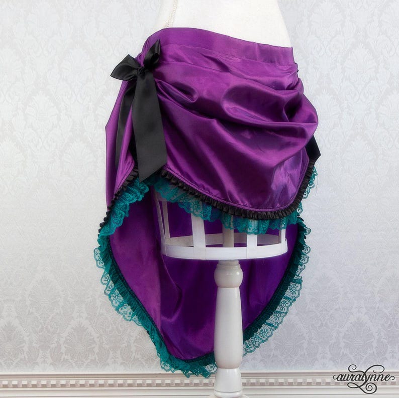 Pu&ograve; includere: Gonna in raso viola con design asimmetrico, con un fiocco nero sul lato. La gonna &egrave; rifinita con pizzo turchese e bordatura nera, creando un effetto a strati. Presentata su un manichino bianco.