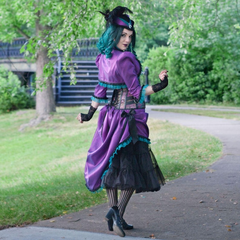 Pu&ograve; includere: Un outfit viola e nero di ispirazione steampunk. L'ensemble comprende una giacca viola con finiture turchesi, un corsetto e una gonna a strati. L'outfit &egrave; accessoriato con un cappello, guanti e calze a righe. Lo sfondo &egrave; un ambiente di parco.