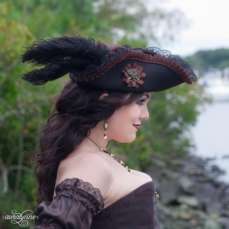 May include: A black pirate hat with brown trim, lace, and a decorative emblem featuring a skull. The hat is adorned with black feathers. The image shows a woman wearing the hat, a brown corset, and a brown sleeve with lace.