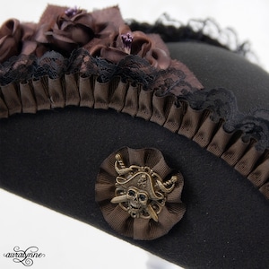 May include: Black pirate hat with brown ribbon and lace trim. The hat is decorated with brown fabric roses and a metal skull and crossbones emblem. A unique accessory for cosplay or themed events.