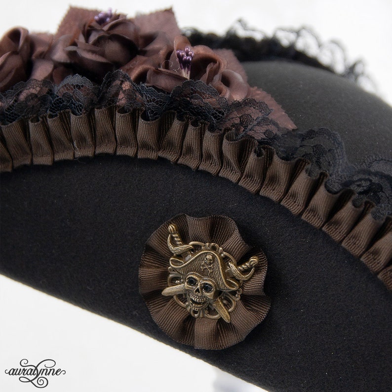 May include: Black pirate hat with brown ribbon and lace trim. The hat is decorated with brown fabric roses and a metal skull and crossbones emblem. A unique accessory for cosplay or themed events.