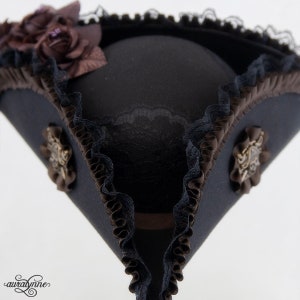 May include: A navy blue tricorn hat with brown and black accents. The hat features decorative brown ribbon trim, black lace edging, and small brown floral embellishments. Two ornate bronze-colored buttons are on the sides.