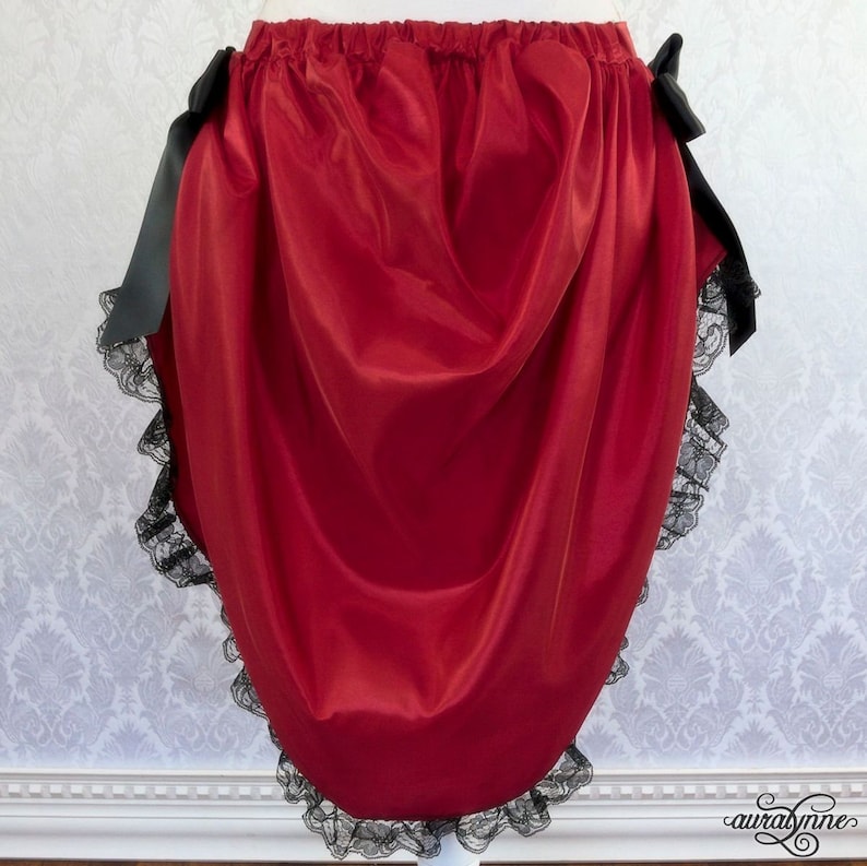K&ouml;nnte beinhalten: Ein roter Satinrock mit schwarzer Spitzenborte und schwarzen Schleifenb&auml;ndern. Der Rock ist in der Taille gerafft und hat eine flie&szlig;ende, drapierende Silhouette.