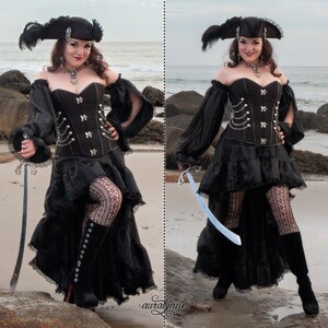 May include: A woman in a black pirate costume with a black corset, a black skirt with ruffles, and black boots. She is holding a sword in each hand and is standing on a beach.