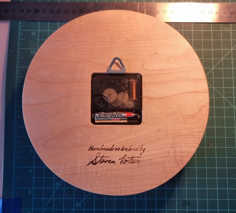 May include: A round wooden clock back with a clock mechanism and an Energizer battery. The text "Handmade in Ireland by Steven Potter" is inscribed on the wood. A metal ruler is visible in the background.