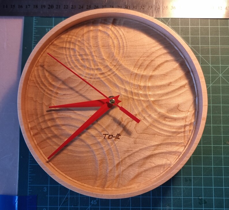 May include: A round wooden clock with a light wood grain finish. The clock features bright red hands and a red second hand. The text "To-it" is printed at the bottom. The clock's diameter is approximately 10 inches.