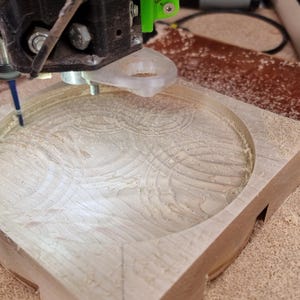 May include: A CNC machine carving a circular design into a light-colored wooden block. The machine's bit is actively removing material, creating a detailed pattern. Sawdust surrounds the work area.
