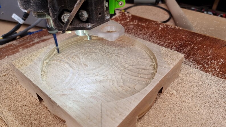 May include: A CNC machine carving a circular design into a light-colored wooden block. The machine's bit is actively removing material, creating a detailed pattern. Sawdust surrounds the work area.