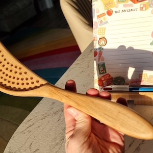 May include: A light brown wooden kitchen utensil with a curved, perforated head and a smooth, tapered handle. The tool features a series of small, evenly spaced circular holes across the wide, crescent-shaped section designed for straining.