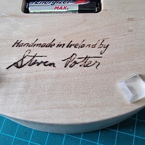 May include: Close-up of the back of a wooden clock. The text "Handmade in Ireland by Steven Potter" is written in black ink. Two clear, square, adhesive bumpers are attached to the wooden surface. An Energizer Max battery is visible.