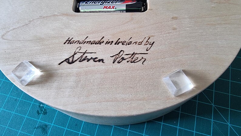 May include: Close-up of the back of a wooden clock. The text "Handmade in Ireland by Steven Potter" is written in black ink. Two clear, square, adhesive bumpers are attached to the wooden surface. An Energizer Max battery is visible.
