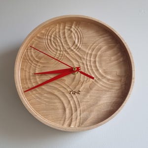 May include: Round wooden wall clock with a light brown finish and red hands. The clock face has a carved, textured design. The text "Ta-it" is printed on the clock face.