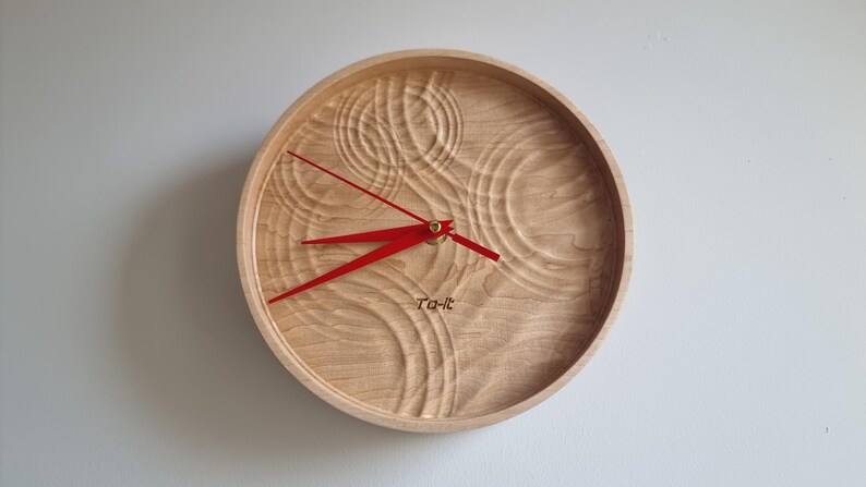 May include: Round wooden wall clock with a light brown finish and red hands. The clock face has a carved, textured design. The text "Ta-it" is printed on the clock face.