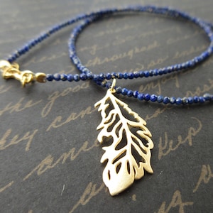 May include: A gold feather pendant necklace with a delicate lapis lazuli beaded chain. The feather pendant is intricately detailed and has a cutout design.