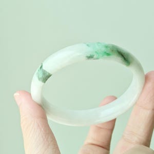 Op de afbeelding: Een gladde, ronde armband gemaakt van gepolijste steen. De armband heeft een doorschijnende witte basiskleur met opvallende, levendig groene minerale insluitingen en vlekken die onregelmatig over het afgeronde oppervlak zijn verdeeld.