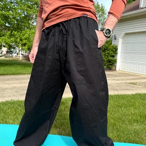 May include: Black, loose-fitting pants with a drawstring waist and pockets. The pants are made of a lightweight fabric and are worn outdoors on a blue mat. The person is wearing a long-sleeved orange shirt.