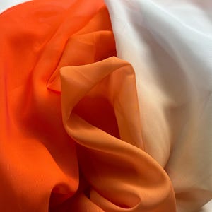 May include: Close-up of layered fabric in shades of orange and white. The fabric is draped and folded, creating a textured effect. The colors blend together, with the orange transitioning to a lighter hue.