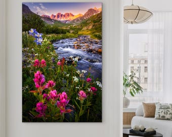 May include: A canvas print of a mountain landscape featuring a flowing river and vibrant wildflowers. The artwork showcases a range of colors, including pink, blue, and green, with a mountain range in the background. The scene is illuminated by the warm glow of the setting sun.