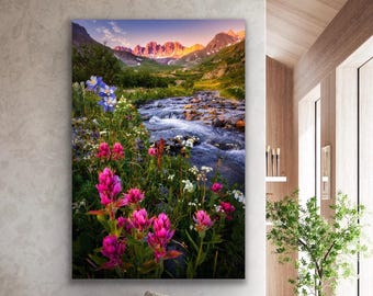 May include: A vertical wall art print featuring a mountain landscape at sunrise. The foreground displays vibrant pink Indian paintbrush flowers and blue columbines. A rocky stream flows through a green valley toward snow-capped mountain peaks.