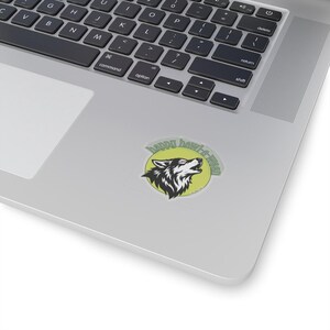 May include: A circular sticker featuring a stylized black and white wolf head howling against a pale yellow circular background. The text "happy howl-o-ween" is printed in a dark green arched font above the wolf's head on the sticker surface.